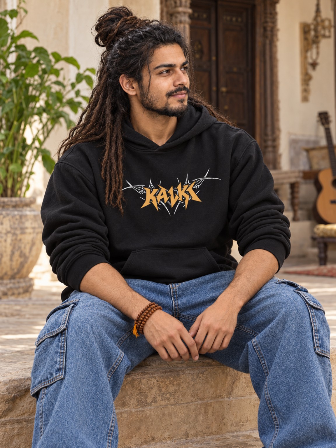 Rudra seated in a courtyard wearing the black Kalki hoodie, with his guitar nearby